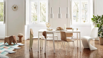A modern dining room with white walls, large windows, a white dining table, mixed wood and white chairs, three pendant lights, plants, decorative items on a wooden floor, and the Vitra Wall Clock - Ball Clock by Vitra for a contemporary accent.