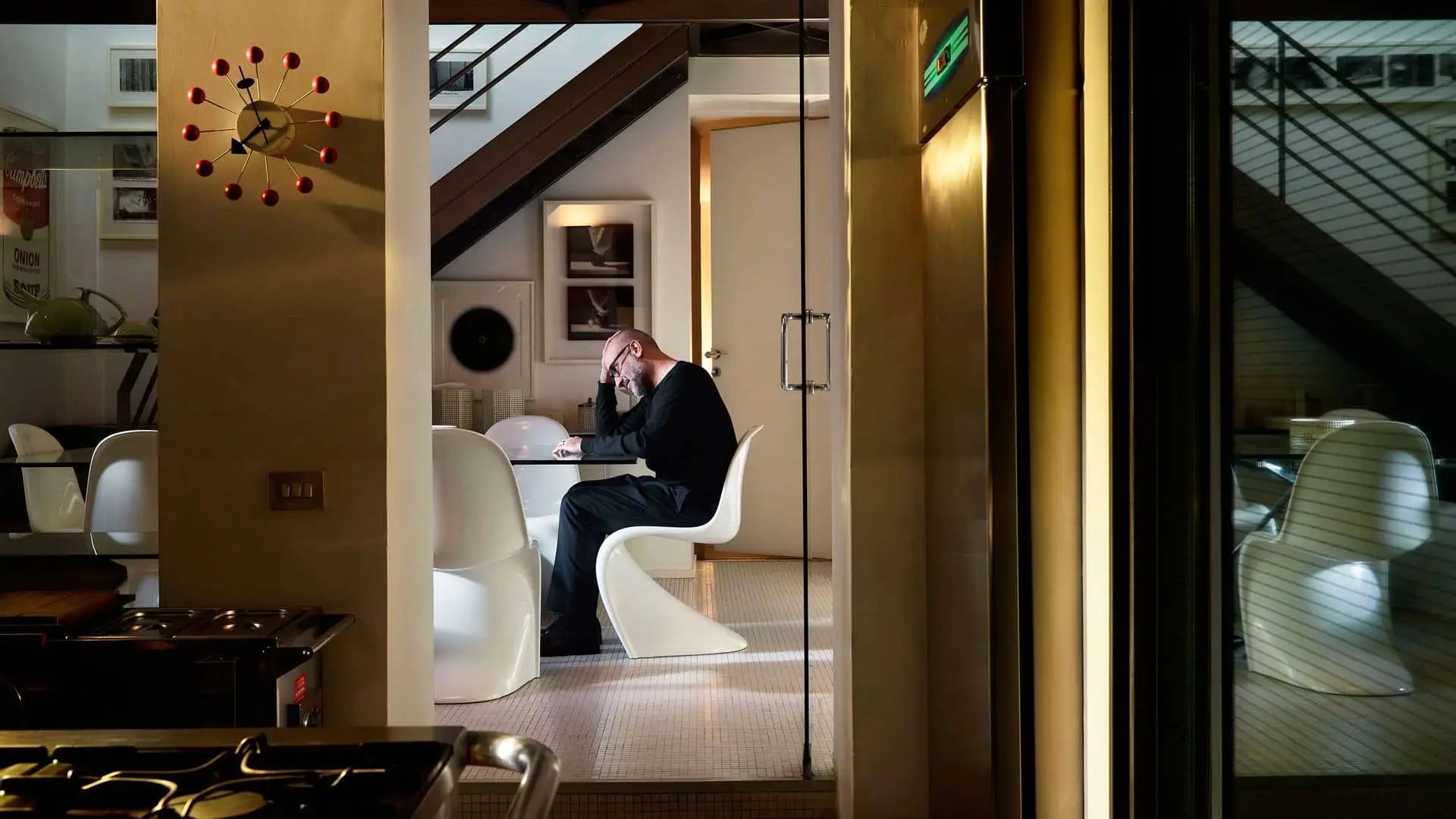A man works on his laptop at a white table in a stylish dining area. The Vitra Wall Clock - Ball Clock by Vitra adds a vibrant, modern accent, while warm lighting highlights the stairs and wall art in the background.