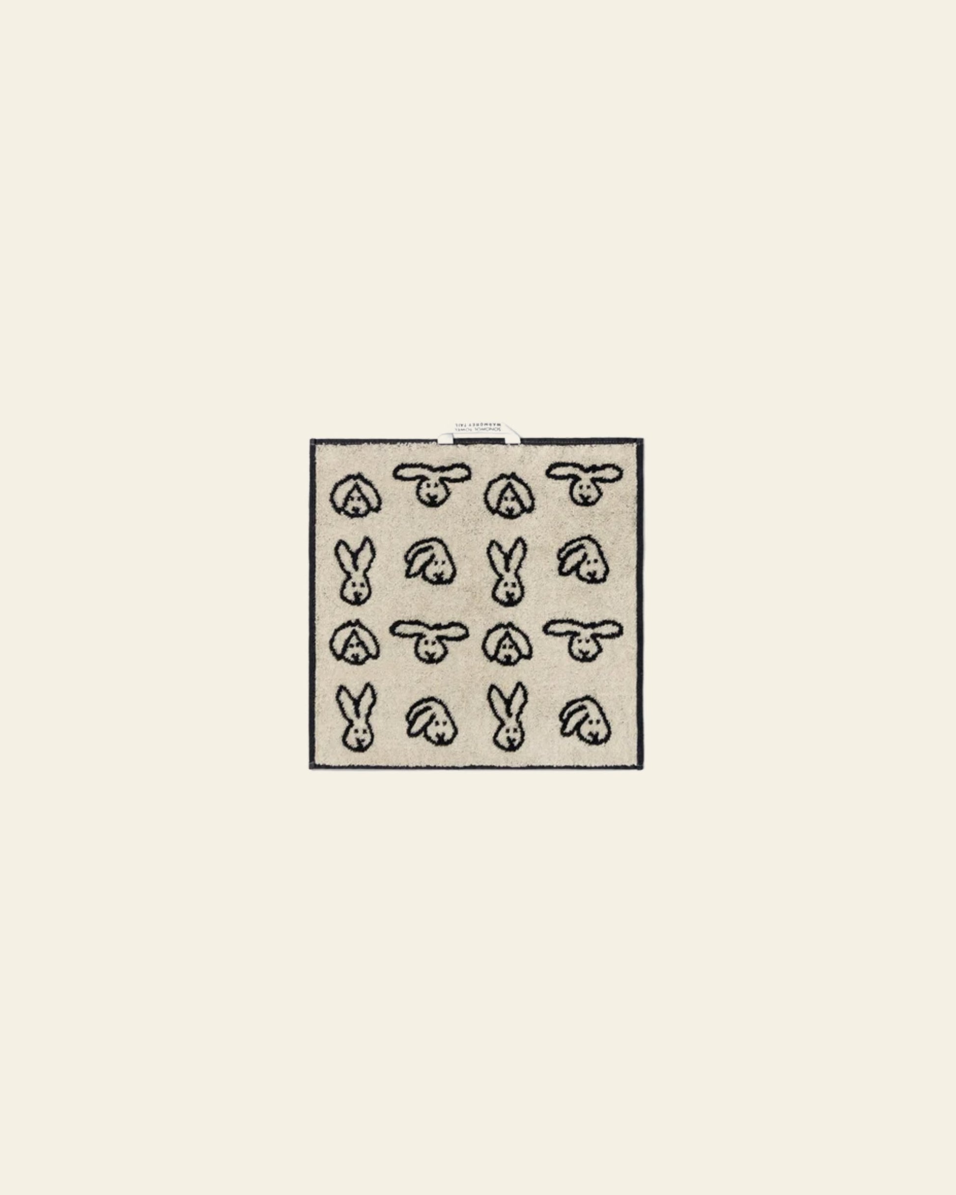 The WARMGREY TAIL Bunny Bunny Hand Towel by WARMGREY TAIL is a square beige cotton towel with a black outline grid of twelve bunnies in various angles on a plain background, plus a small white tag at the top center.