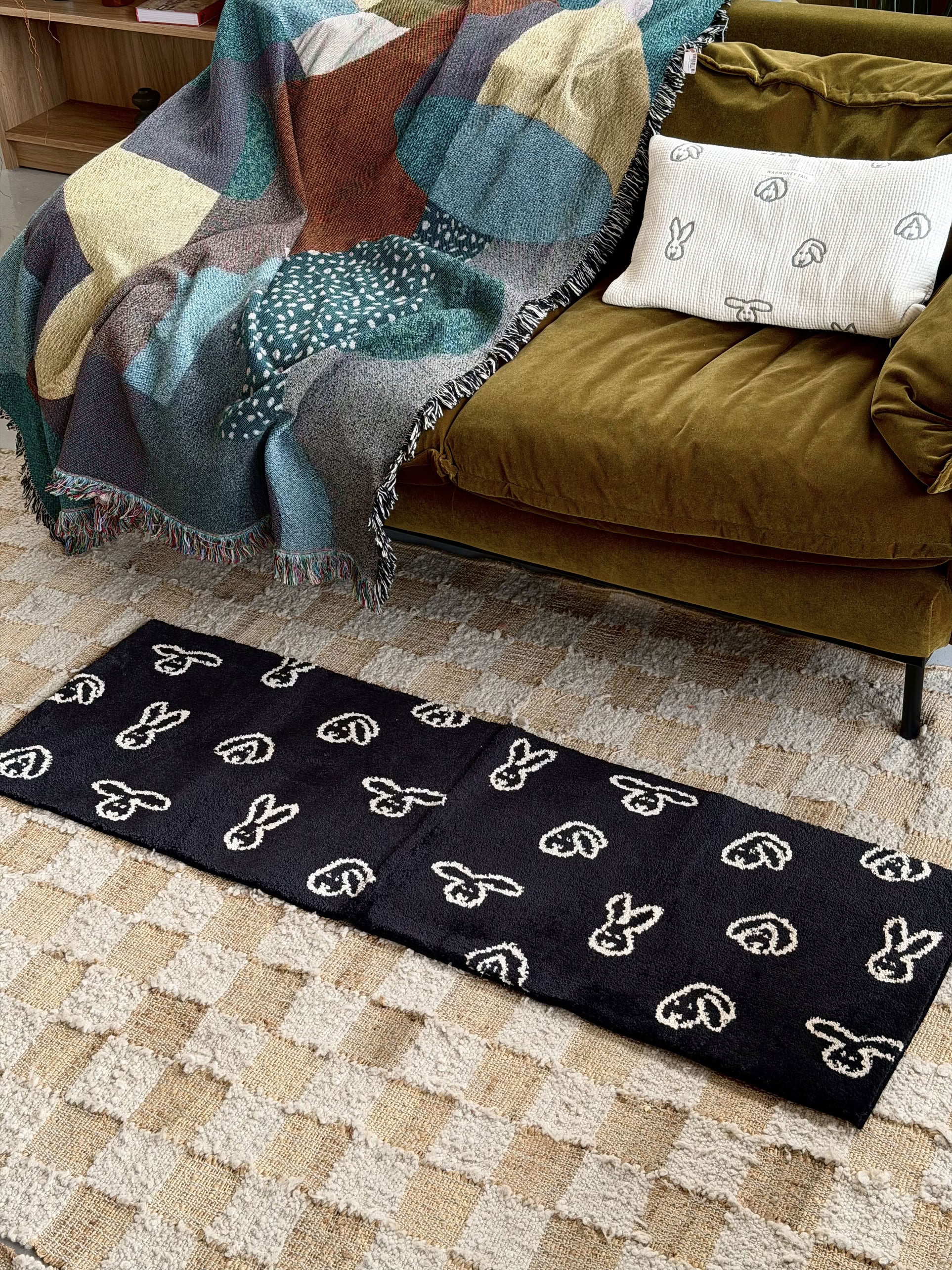 A WARMGREY TAIL Bunny Bunny Rug (140x45) in black, featuring white-outlined bunny and heart motifs, is laid over a beige checkered carpet in front of a green velvet sofa adorned with a patterned throw and a white pillow decorated with bunny designs.