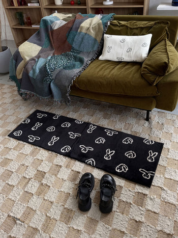 A green couch adorned with a patterned blanket and a bunny pillow sits in a cozy living room, where the WARMGREY TAIL Bunny Bunny Rug (140x45) - Black, featuring jacquard-woven white bunny outlines on black, lies nearby along with black shoes resting on a beige checkered carpet.