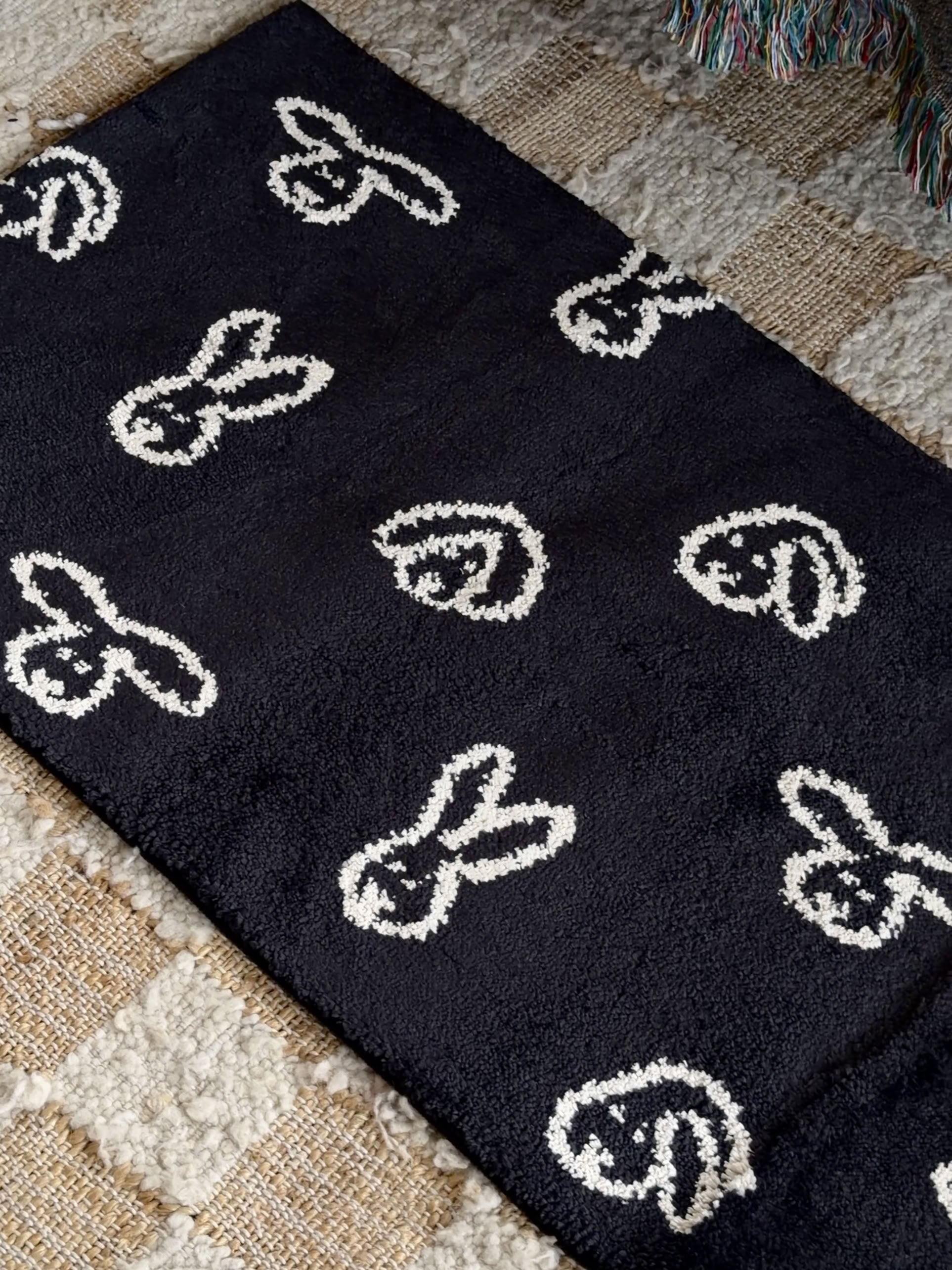The WARMGREY TAIL Bunny Bunny Rug (140x45) - Black, featuring simple white bunny face patterns, is laid on a beige, textured, checkered carpet. This jacquard-woven rug from WARMGREY TAIL adds a playful touch to the space, while a colorful fringed throw is partially visible at the top right corner.