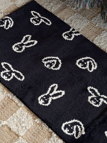 The WARMGREY TAIL Bunny Bunny Rug (140x45) - Black, featuring simple white bunny face patterns, is laid on a beige, textured, checkered carpet. This jacquard-woven rug from WARMGREY TAIL adds a playful touch to the space, while a colorful fringed throw is partially visible at the top right corner.