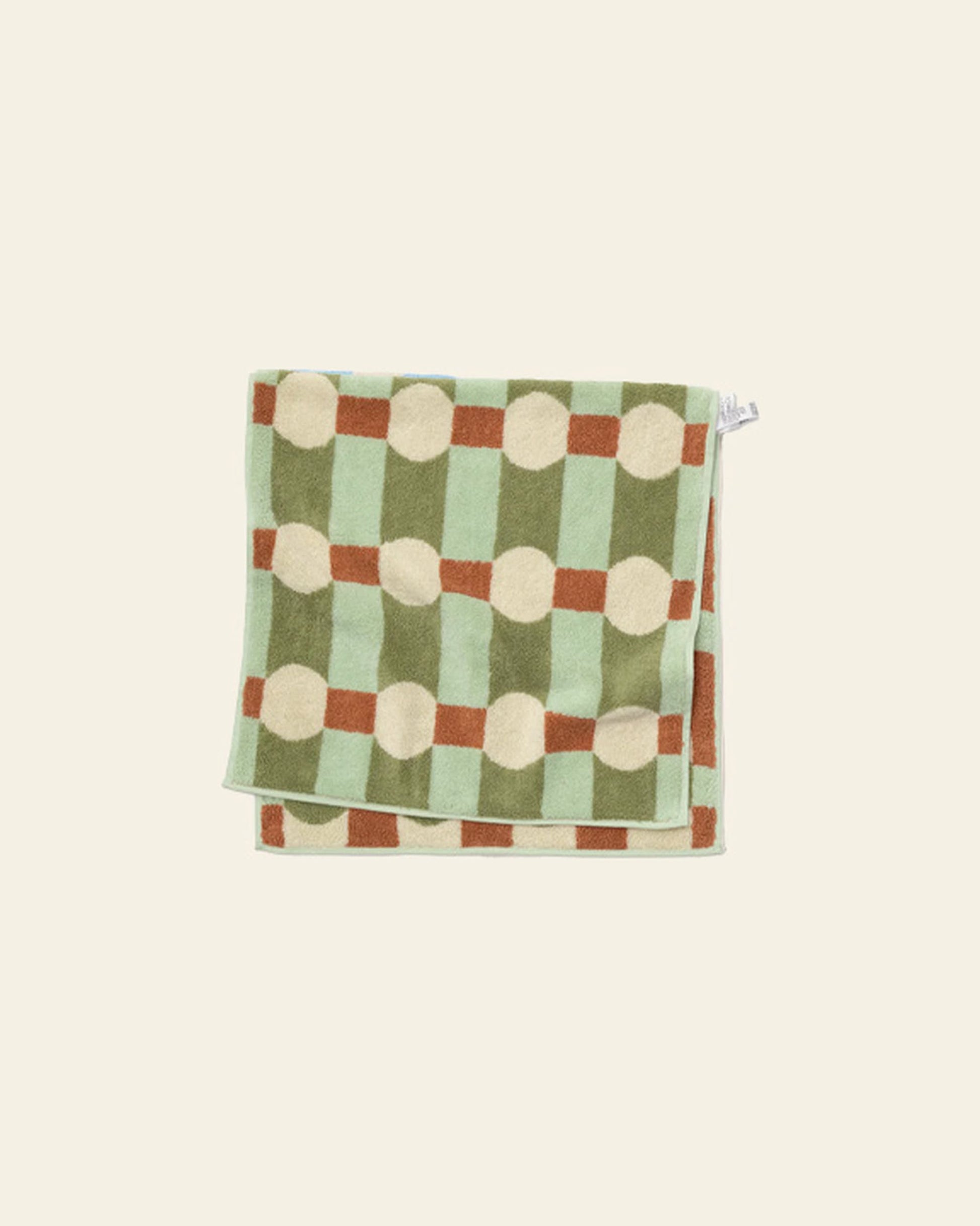 The WARMGREY TAIL Dot Check Face Towel by WARMGREY TAIL is a folded cotton terry towel with a geometric green, brown, and cream squares and circles pattern set against an off-white background.