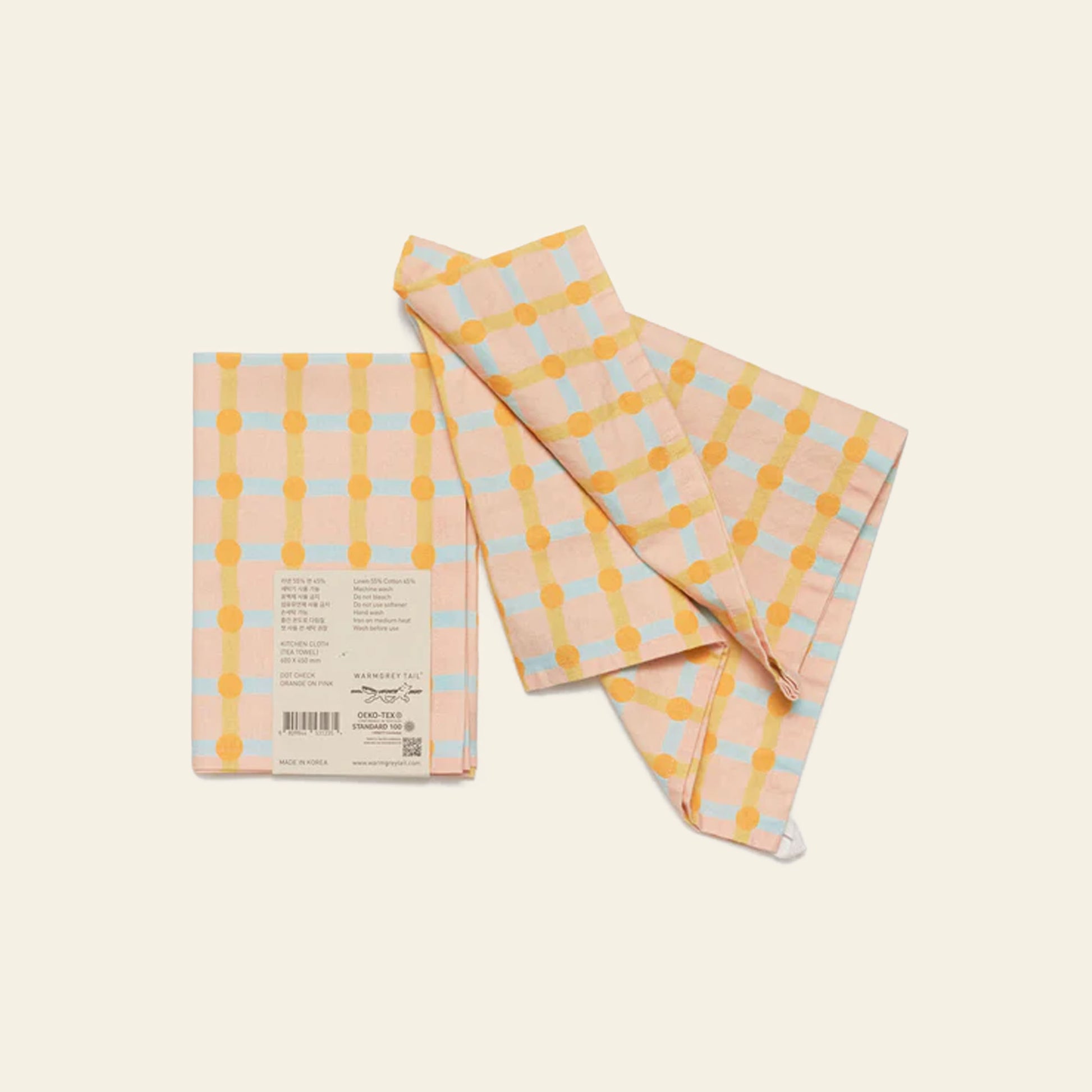 Two WARMGREY TAIL Dot Check Kitchen Cloths in pastel orange and yellow are displayed on a light background—one folded with a label and the other loosely draped.