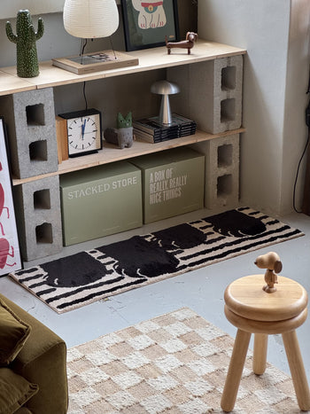 A cozy room corner features a cinder block and wood shelf with books, art, a lamp, clock, and plant. Beside a wooden stool lies the WARMGREY TAIL Paw Rug (140x45) by WARMGREY TAIL, showcasing a zebra pattern and non-slip grip.