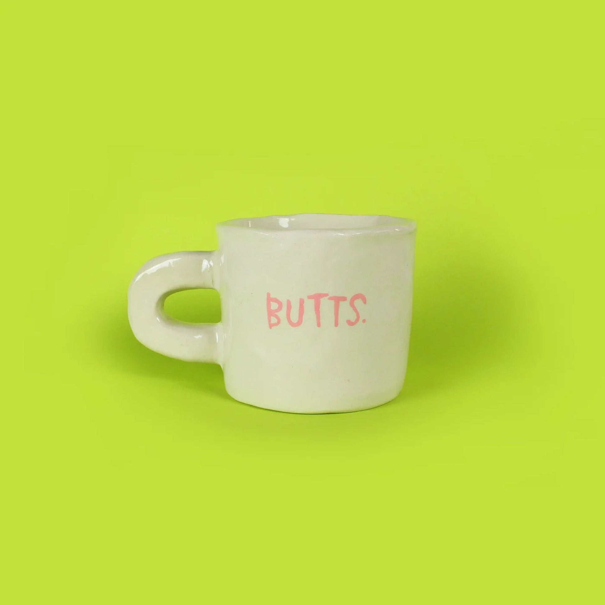 The Wobbly Studio Butts Mug by Wobbly Studio features BUTTS. in pink on white ceramic, set against a bright lime green background—a playful and humorous addition to any collection or fun conversation starter.