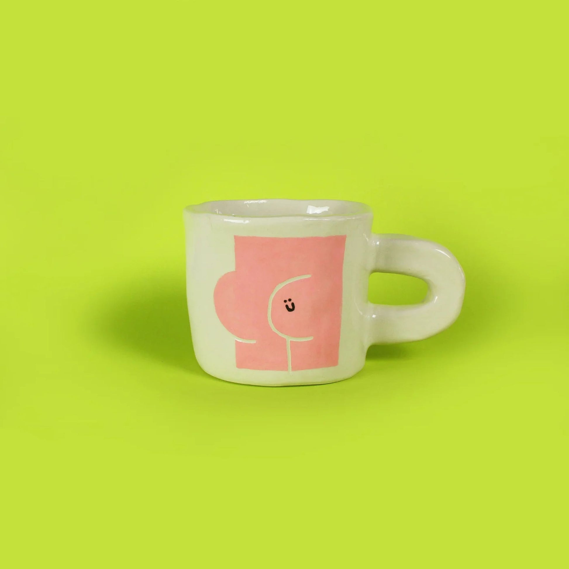 The Wobbly Studio Butts Mug by Wobbly Studio is a white ceramic mug with a large handle, set against a green background. Its pink abstract design and playful smiling face make it a perfect conversation starter.