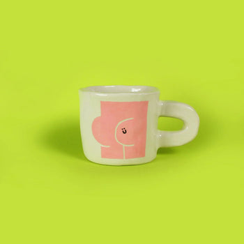 The Wobbly Studio Butts Mug by Wobbly Studio is a white ceramic mug with a large handle, set against a green background. Its pink abstract design and playful smiling face make it a perfect conversation starter.