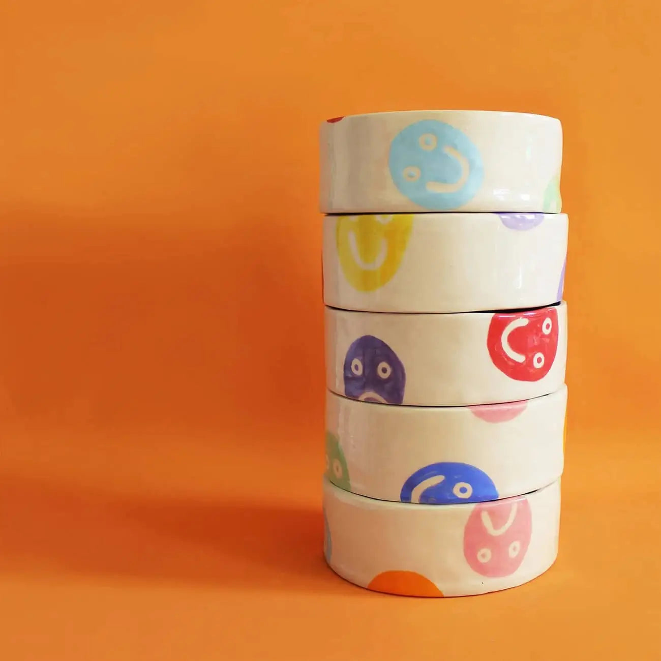 A stack of five handcrafted white Wobbly Studio Doggo Bowl, Bigger by Wobbly Studio, each with multicolour smiley faces, set against an orange background. A cheerful kitchen addition.
