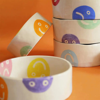 Five Wobbly Studio Doggo Bowl, Bigger ceramic bowls with multicolour smiley and frownie faces—yellow, purple, pink, green, blue—stacked on orange. Playful design by Wobbly Studio to brighten up any space or use as a pet bowl.