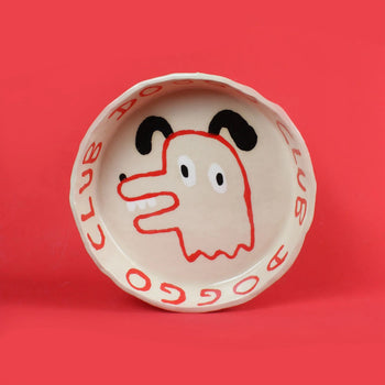 The Wobbly Studio Doggo Club Ti Chat Bowl is a ceramic plate featuring a cute dog face in red, black, and cream. “DOGGO CLUB” repeats on the rim in red over cream with a bold red background—perfect for your pet’s meals.