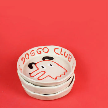 Three ceramic bowls are stacked on a red background. The top bowl is the Wobbly Studio Doggo Club Ti Chat Bowl by Wobbly Studio, featuring a cartoon dog face and DOGGO CLUB lettering—cute and functional for your pet’s meals.