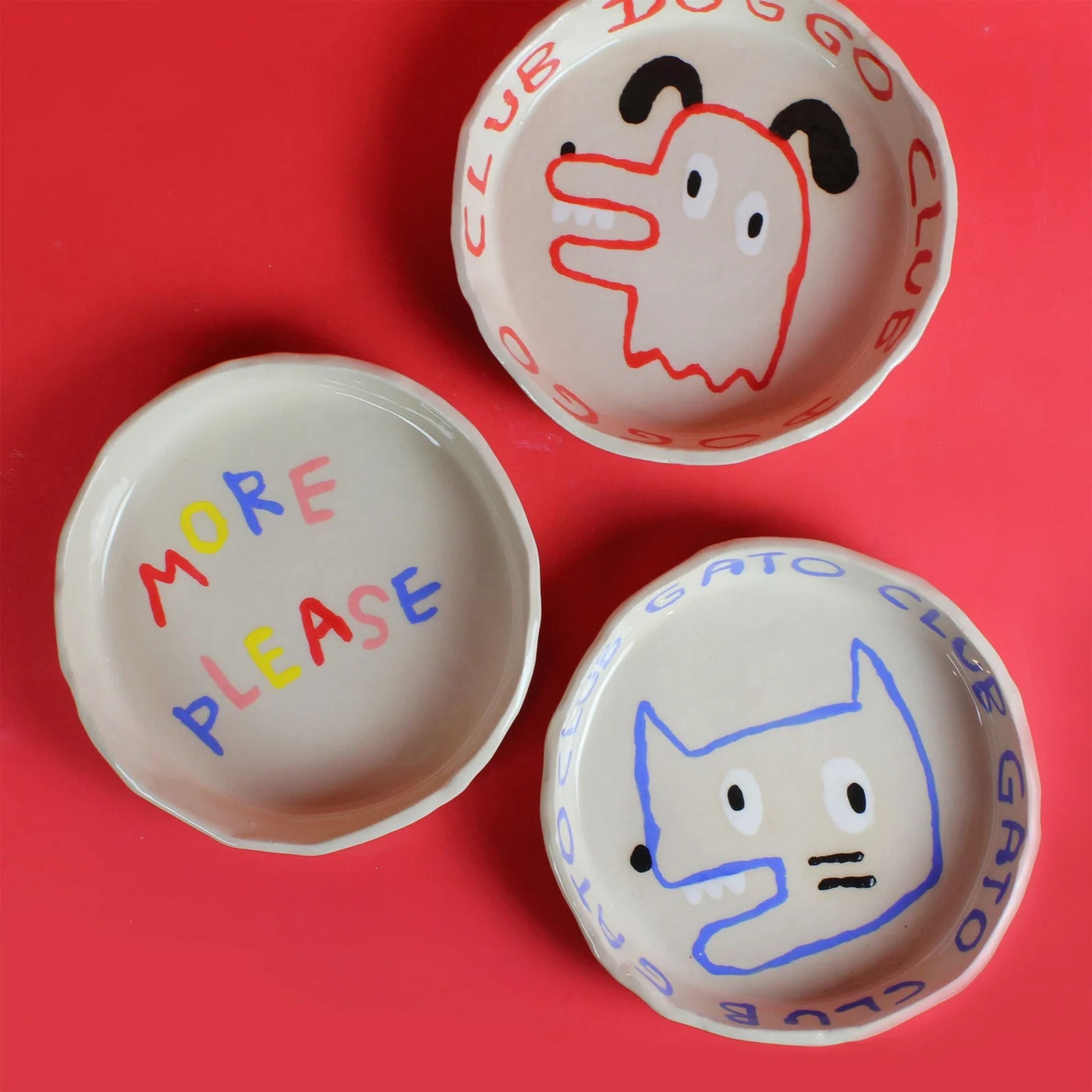 Three handmade Wobbly Studio ceramic bowls sit on a red surface: one says “MORE PLEASE,” one is the Doggo Club Bowl with a cartoon pup, and the third is the Wobbly Studio Doggo Club Ti Chat Bowl for your pet’s meals.