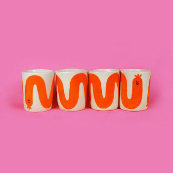 Four Wobbly Studio Happy Saucisse Tumblers, handmade earthenware cups with orange snake-like designs and quirky faces, are lined up against a bright pink background.