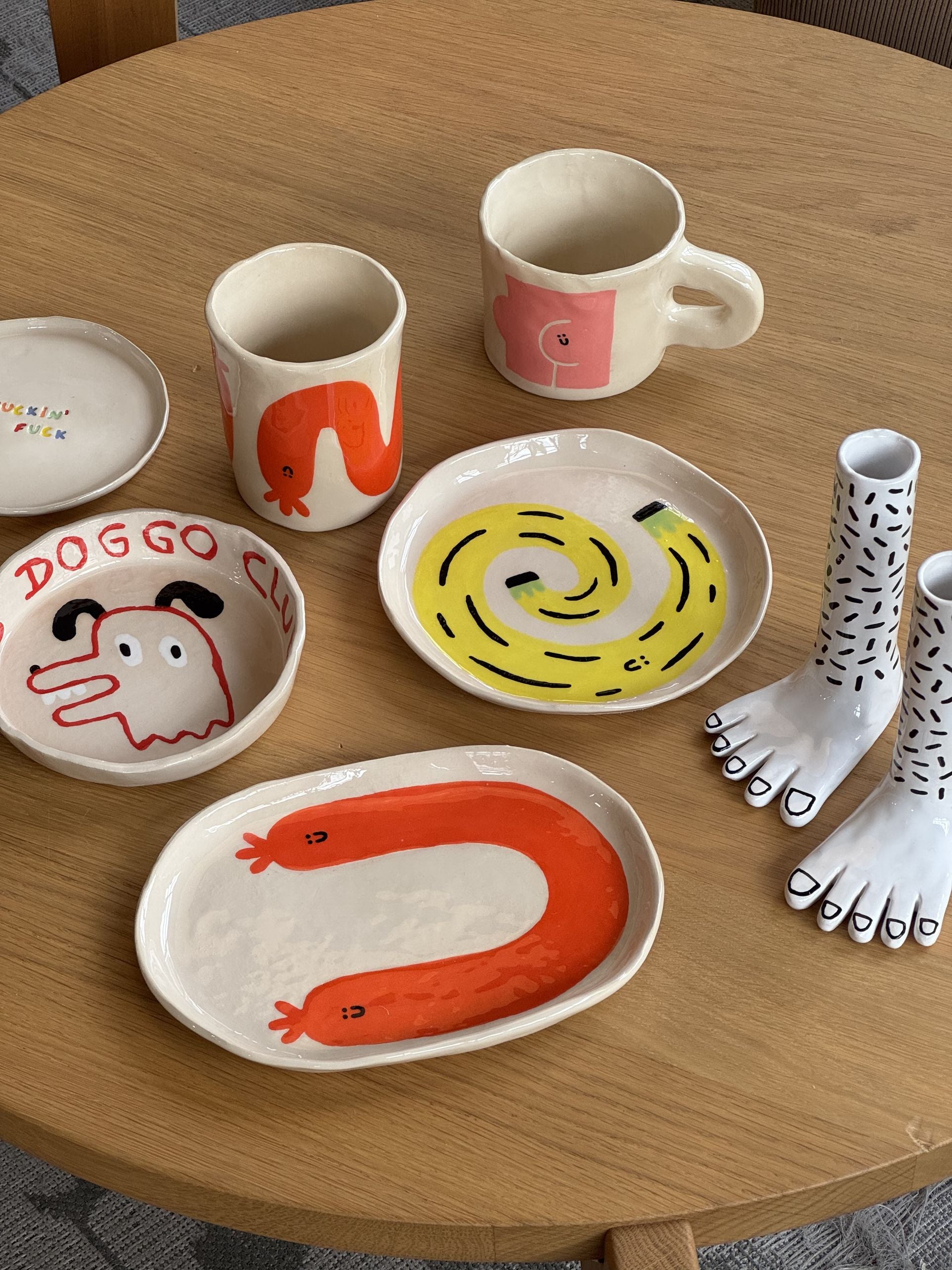A set of quirky ceramic dishes and cups from Wobbly Studio, including the Happy Saucisse Tumbler, featuring playful designs like dog faces, abstract snakes, tall patterned feet, and unique illustrations on a round wooden table.