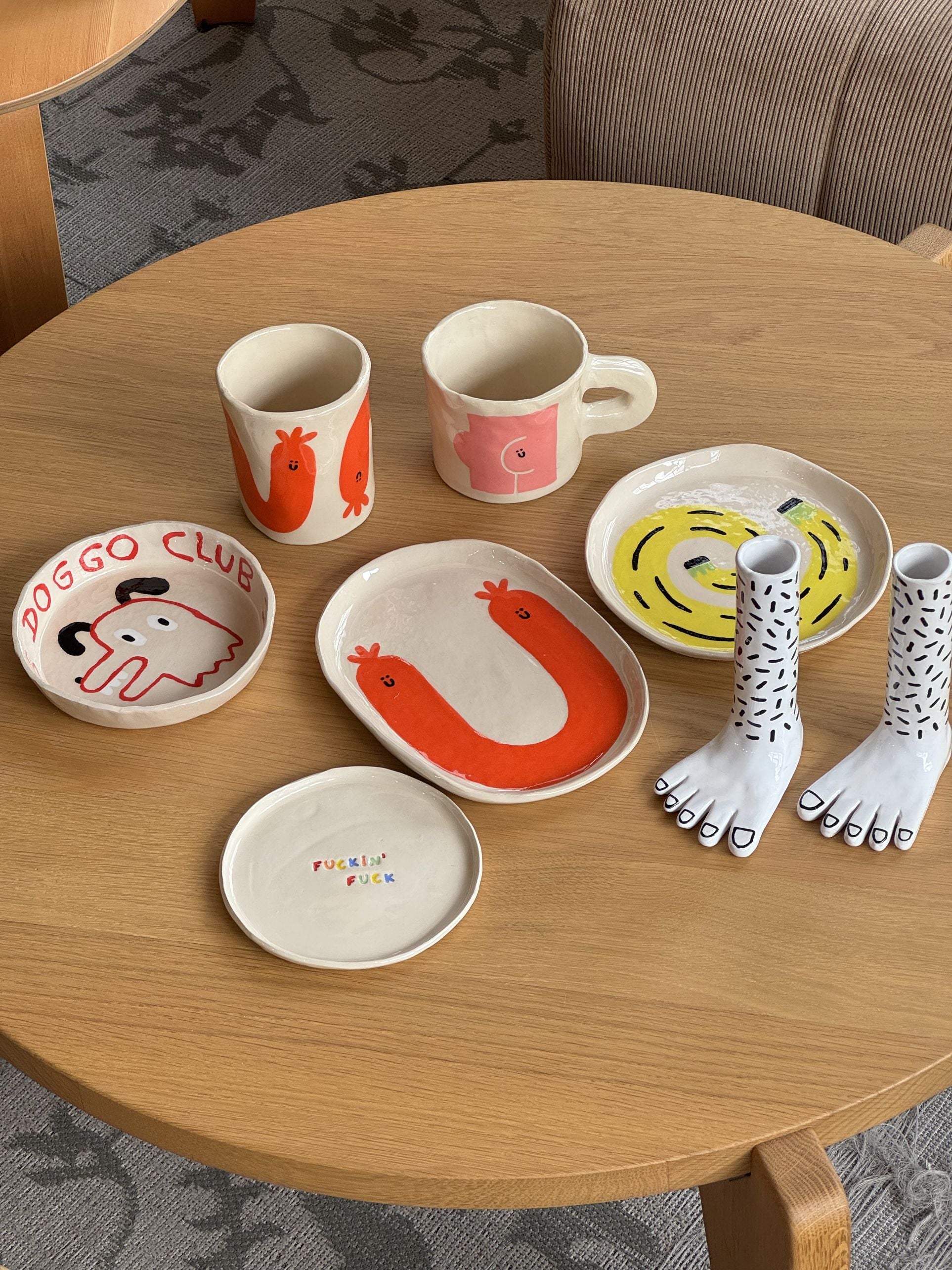 A wooden table displays Wobbly Studios handmade earthenware dishes, mugs, foot-shaped candle holders, and the playful Happy Saucisse Tumbler with vibrant animal and foot illustrations plus quirky text like “DOGGO CLUB.”.