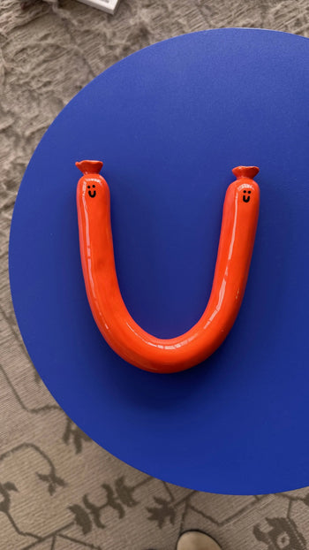 The Wobbly Studio Happy Saucisse Wall Decor by Wobbly Studio is a U-shaped ceramic sculpture with red smiling faces on both ends, set on a blue round base—perfect for adding quirky, handcrafted charm to your space.