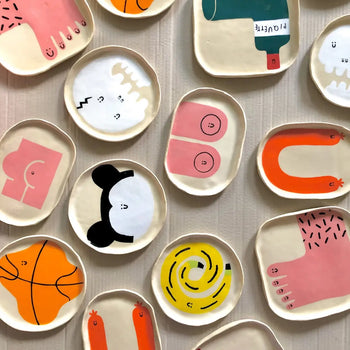 The Wobbly Studio Happy Things Plate - Banane features playful, whimsical designs with bold lines and colors, including cheerful abstract faces, feet, hands, and geometric shapes, arranged on a light textured surface.