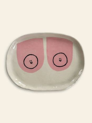 The Wobbly Studio Happy Things Plate - Nénés is a white oval plate with playful designs—two pink semicircles, each with a black circle and smiling face—set on an off-white background.