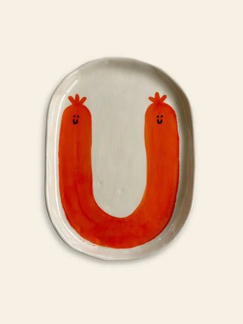 The Wobbly Studio Happy Things Plate - Saucisse is a white ceramic plate featuring a large orange U-shape with smiling faces and crowns, resembling playful sausages in vibrant colors.