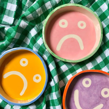 Three Wobbly Studio Mood Trays—round and featuring sad face designs in pink, yellow, and purple—by Wobbly Studio are displayed on a green and white checkered cloth, adding a touch of vibrant colour.