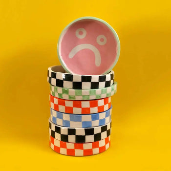 The Wobbly Studio Mood Tray by Wobbly Studio features a stack of four ceramic bowls with colorful checkerboard patterns and a pink bowl with a white sad face, set against a vivid yellow backdrop.