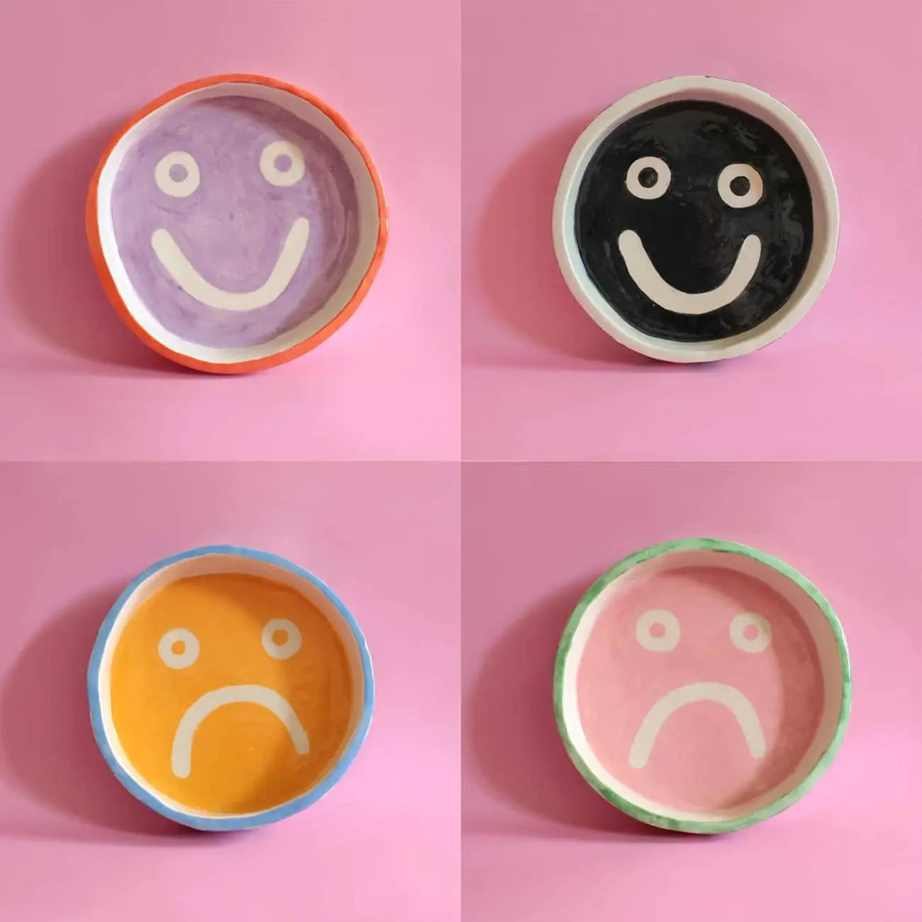 The Wobbly Studio Mood Tray by Wobbly Studio features four ceramic bowls—purple and black with happy faces on top, yellow and pink with sad faces below—arranged on a pink background for a playful, personalized display.