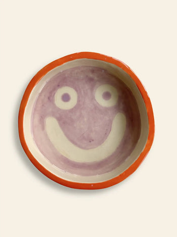 The Wobbly Studio Mood Tray features a round ceramic bowl with a red rim and a cheerful smiling face in light purple on cream, ideal for brightening up personalized displays.