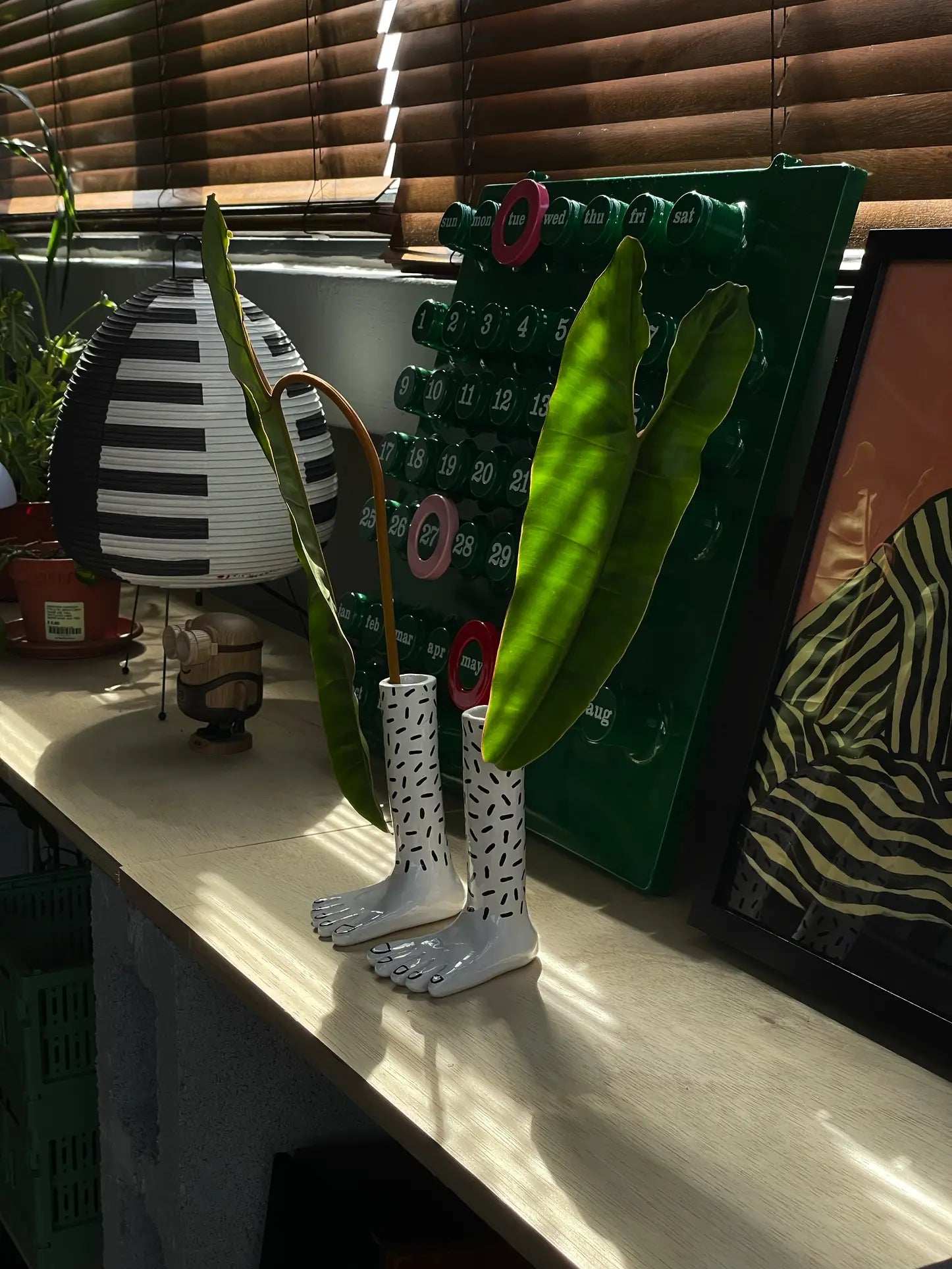A sunlit shelf displays the Wobbly Studio Pair Of Legs Vase by Wobbly Studio, shaped like feet and filled with tall green leaves, alongside a modern lamp, a green wall calendar, potted plants, and framed zebra-striped artwork.