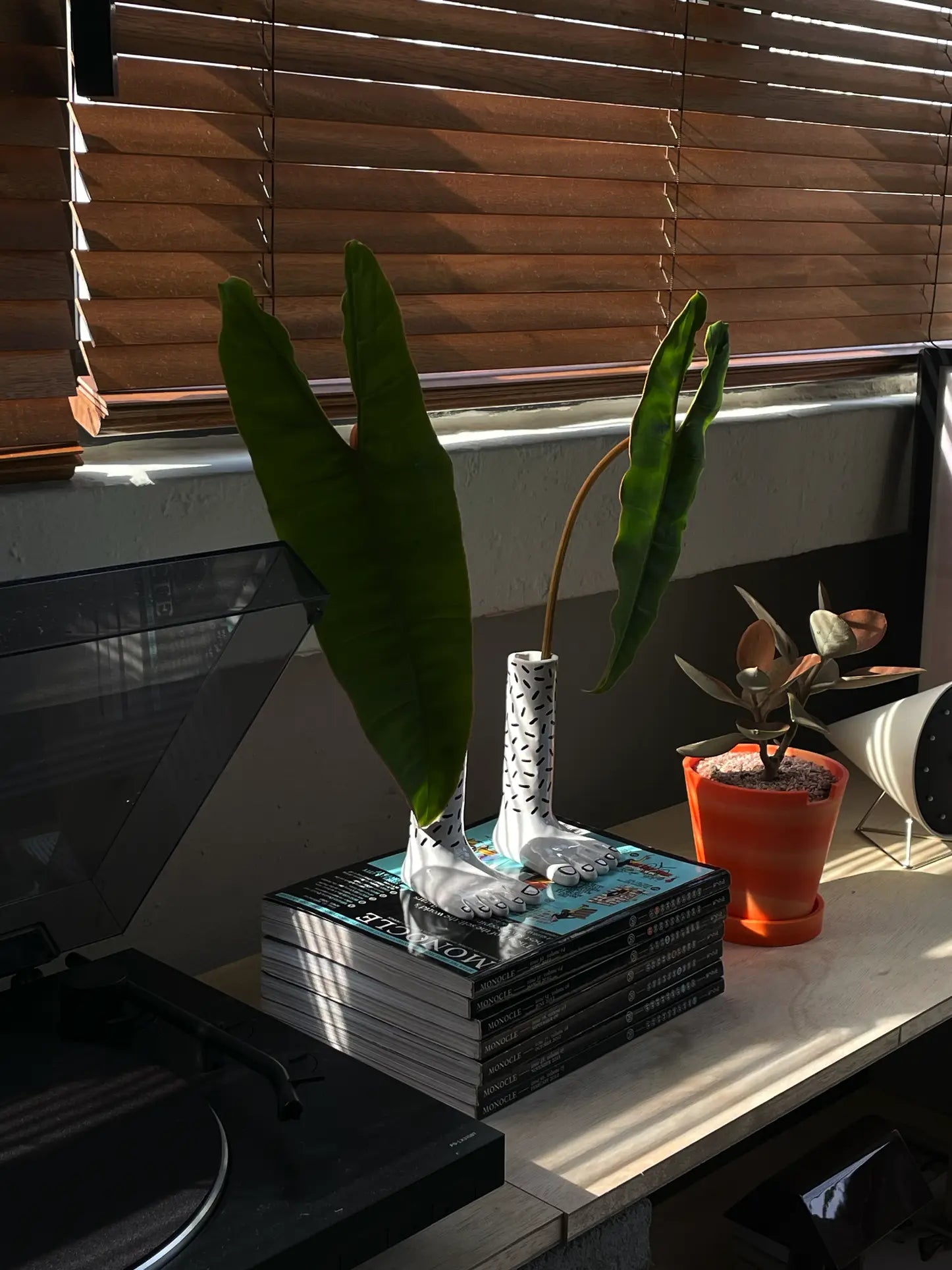 The Wobbly Studio Pair Of Legs Vase by Wobbly Studio, shaped like two feet, displays green leaves on a stack of magazines beside an orange potted plant on a wooden surface with sunlight streaming through window blinds.