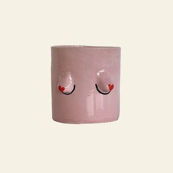 The Wobbly Studio Regular Boobies Pot is a pink handcrafted cylindrical planter with two raised nipple-like shapes, black curved lines, and small red hearts—a playful decor piece for any space by Wobbly Studio.