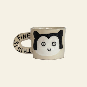 The Wobbly Studio This Is Fine Mug by Wobbly Studio features a beige, handcrafted design with a cartoon animal face and black ears. THIS IS FINE is printed on the handle, adding whimsy to any drinkware collection. Inspired by Marie Chanteur’s illustration.