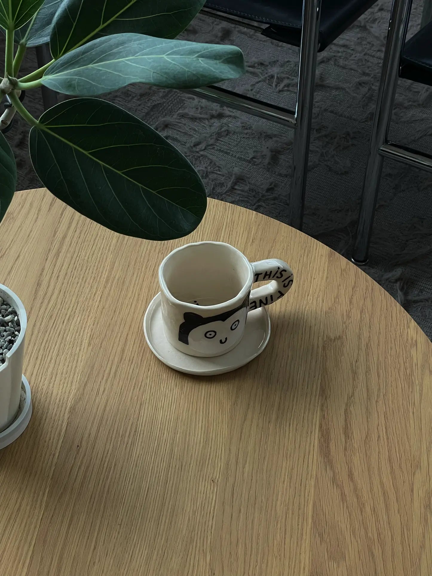 The Wobbly Studio This Is Fine Mug, featuring a cartoon face and handle text, rests on a saucer atop a light wood table beside a leafy potted plant; chairs are visible on the carpeted floor in the background.