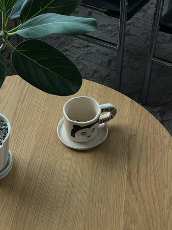 The Wobbly Studio This Is Fine Mug, featuring a cartoon face and handle text, rests on a saucer atop a light wood table beside a leafy potted plant; chairs are visible on the carpeted floor in the background.