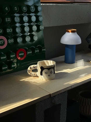 The Wobbly Studio This Is Fine Mug, featuring a cartoon face and ITS FINE on the handle, rests on a sunlit wooden desk alongside a modern lamp and a calendar open from April to July.