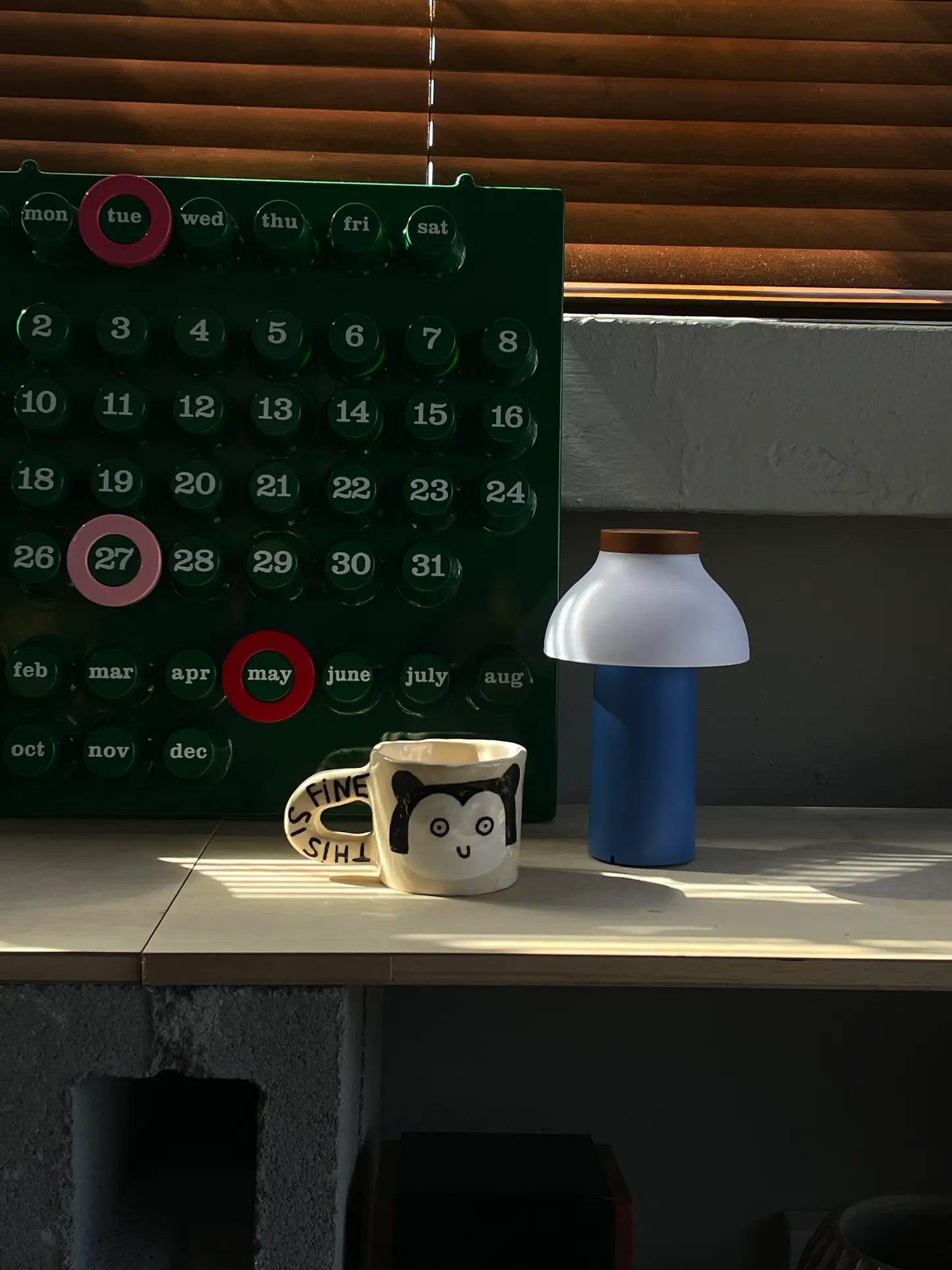 A Wobbly Studio This Is Fine Mug featuring a cartoon panda and ITS FINE sits by a lamp. Behind them is a green reusable calendar with days and months, and window blinds filter in sunlight.