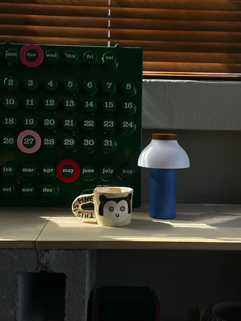 A Wobbly Studio This Is Fine Mug featuring a cartoon panda and ITS FINE sits by a lamp. Behind them is a green reusable calendar with days and months, and window blinds filter in sunlight.