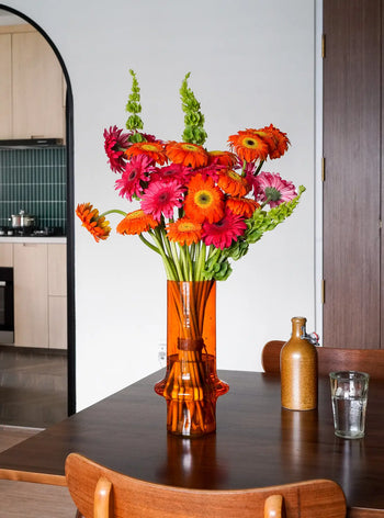 A tall amber glass vase from Wonderland Botanicals Paprika - Wonderland Botanicals Collaboration, filled with a vibrant floral arrangement, stands as a stunning dining table centerpiece amidst a modern kitchen setting.