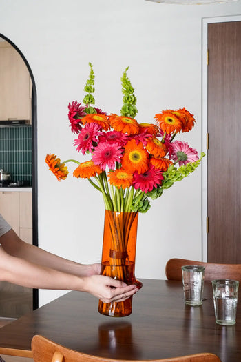 A person places the Wonderland Botanicals Paprika vase, filled with vibrant flowers, on a wooden dining table. Two empty glasses sit nearby, making this showpiece pop against the modern kitchen backdrop.