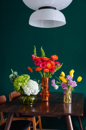 A dark wooden dining table displays the vibrant Paprika - Wonderland Botanicals Collaboration floral arrangement by Wonderland Botanicals, with mixed blooms set against a dark green wall beneath a white hanging lamp. Urban Nature Culture inspired.