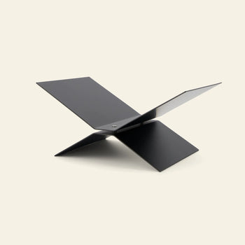 The baebsy Atlas Bookstand by baebsy is a modern, minimalist black metal magazine rack with an X-shaped levitation design and two intersecting flat panels, shown against a light beige background.