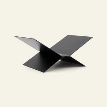 The baebsy Atlas Bookstand by baebsy is a minimalist, black stand with an X-shaped metal frame and four flat panels intersecting at the center for a levitating look, displayed against a light beige background.