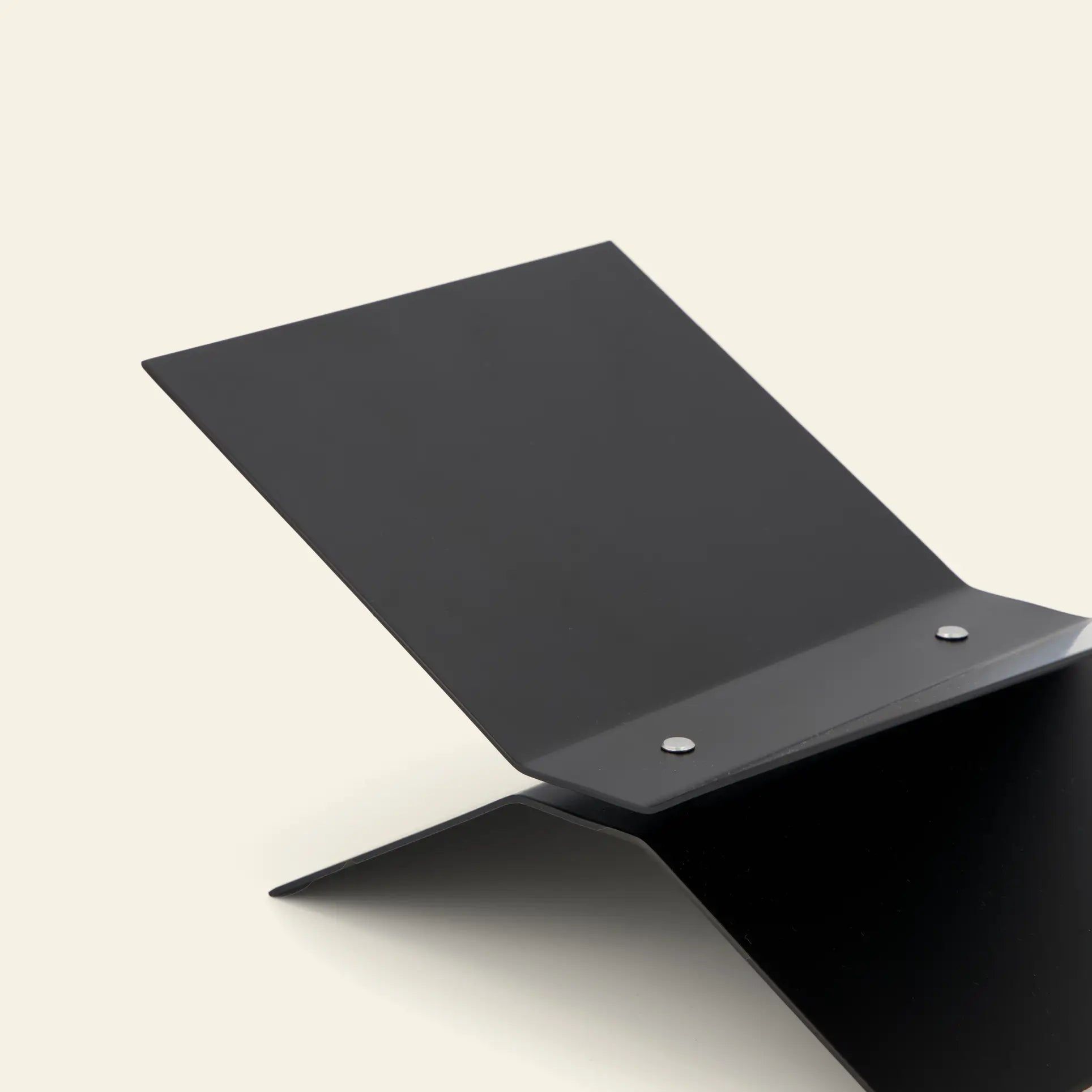 The baebsy Atlas Bookstand by baebsy features a minimalist black metal design with intersecting flat panels and two silver rivets, elegantly displayed on a light beige background. Its levitating form enhances any space.