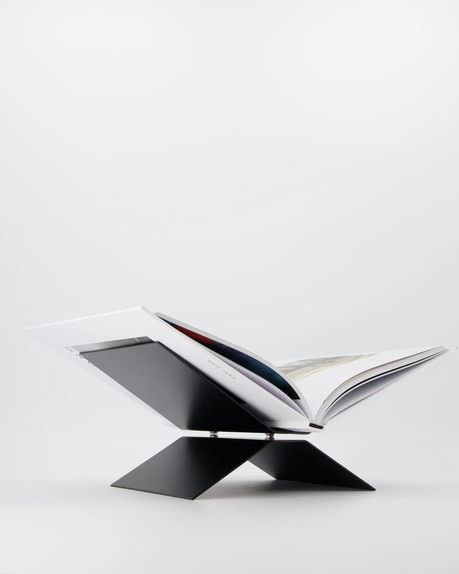 An open book sits on the baebsy Atlas Bookstand by baebsy, a modern black X-shaped stand with a levitation design that elevates and angles books for comfortable reading against a plain white background.