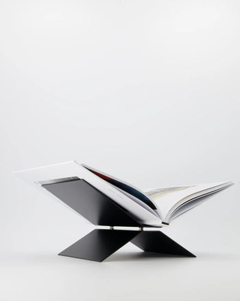 An open book sits on the baebsy Atlas Bookstand by baebsy, a modern black X-shaped stand with a levitation design that elevates and angles books for comfortable reading against a plain white background.