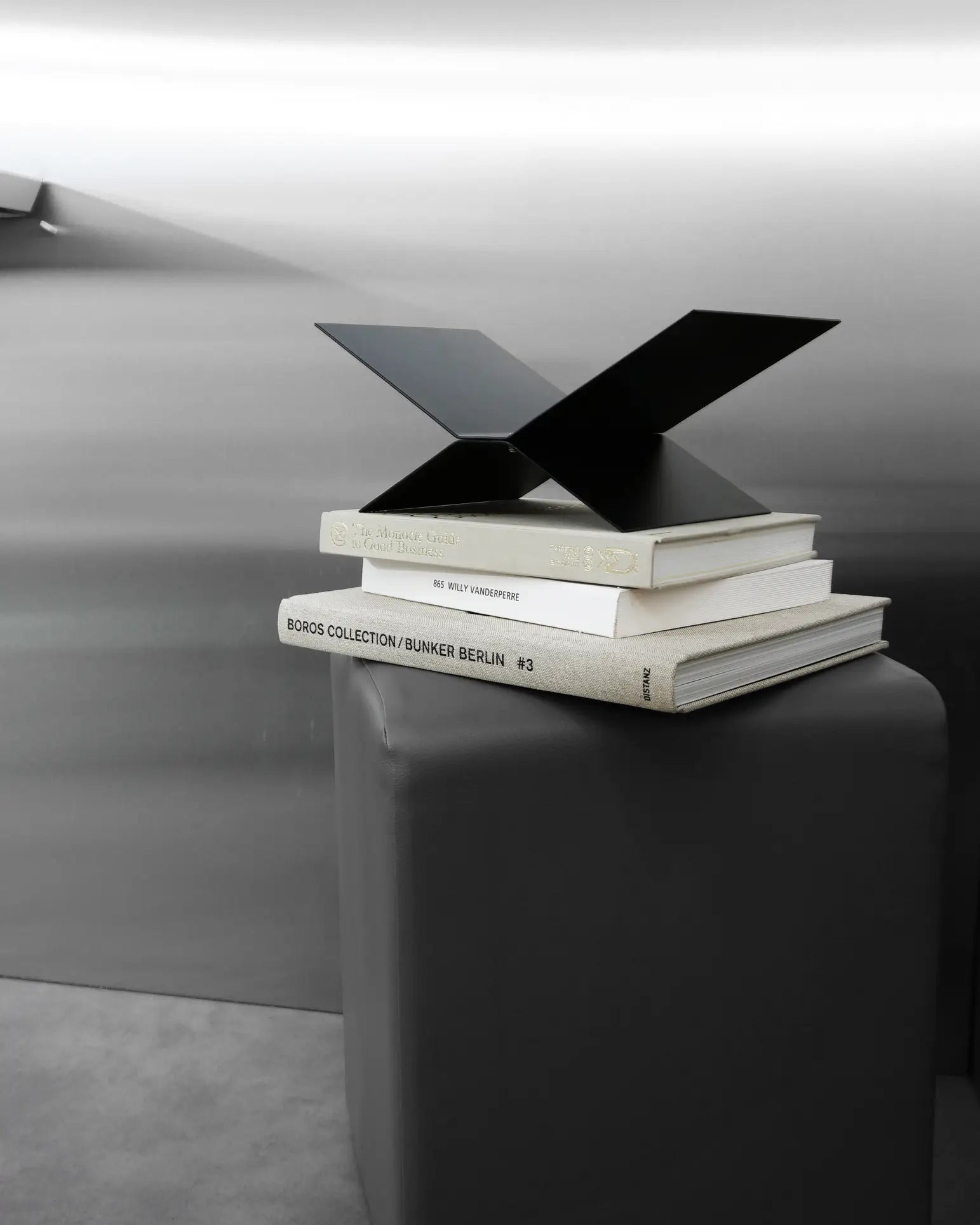 The baebsy Atlas Bookstand by baebsy features a modern black metal sculpture that seems to float above three art books on a gray pedestal, highlighting its sleek levitation design in a minimalist, metallic-toned setting.