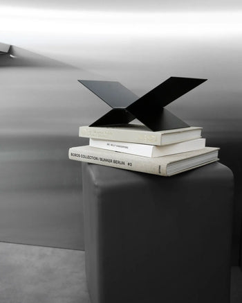 The baebsy Atlas Bookstand by baebsy features a modern black metal sculpture that seems to float above three art books on a gray pedestal, highlighting its sleek levitation design in a minimalist, metallic-toned setting.