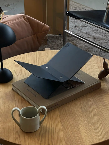 A modern workspace showcases a baebsy Atlas Bookstand holding a large book on a wooden table, alongside a black folder, ceramic mug, decorative piece, and sleek desk lamp, complemented by a cozy chair in the background.