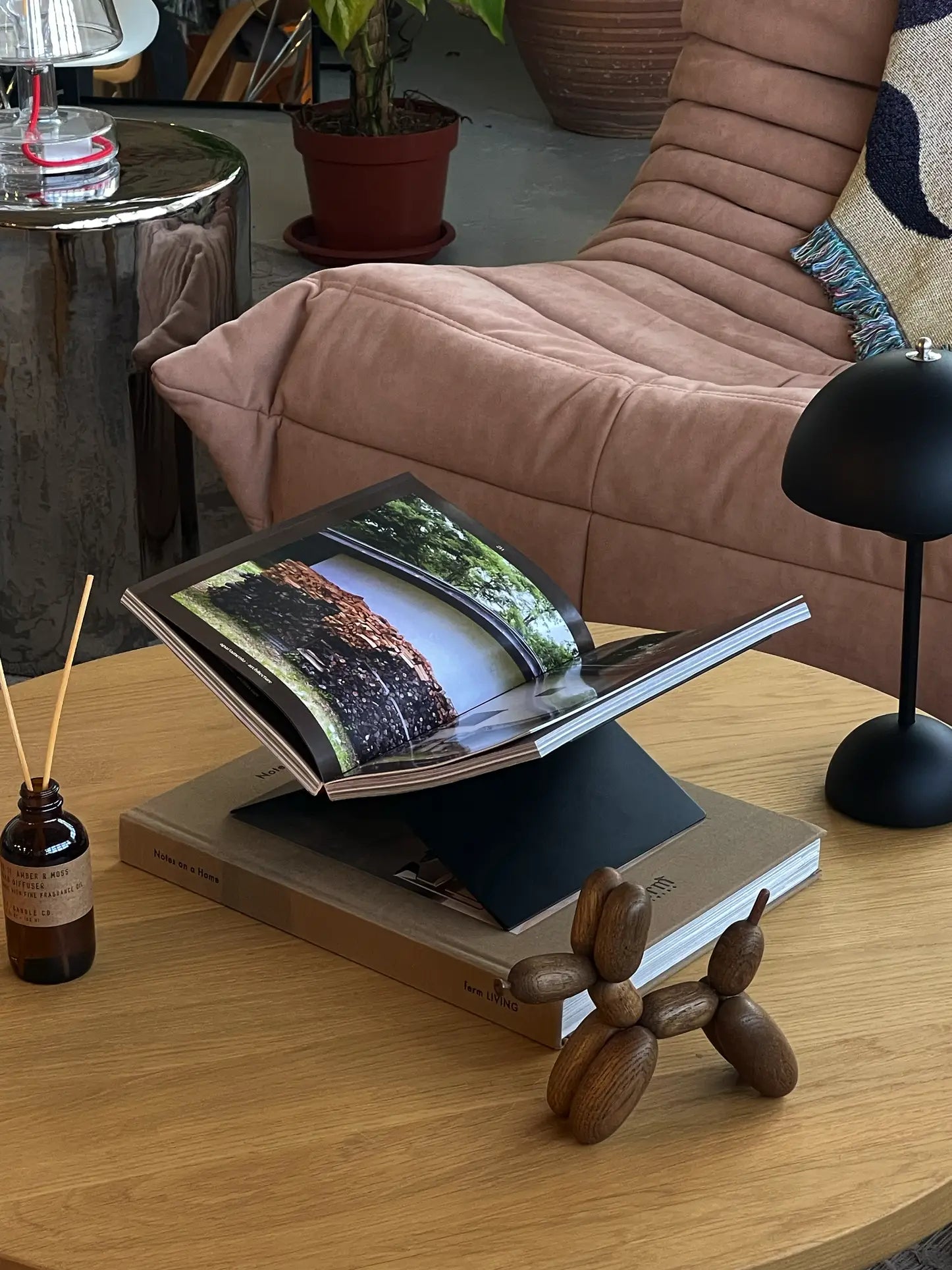 A coffee table features a wooden dog figurine, the baebsy Atlas Bookstand displaying an open photography book, a black lamp, a reed diffuser, and a brown couch with a blanket in the background.